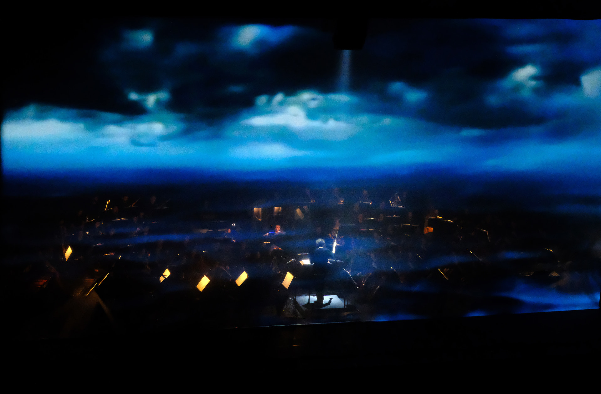 Orchestra playing beneath a vast projection of moving cloudscapes